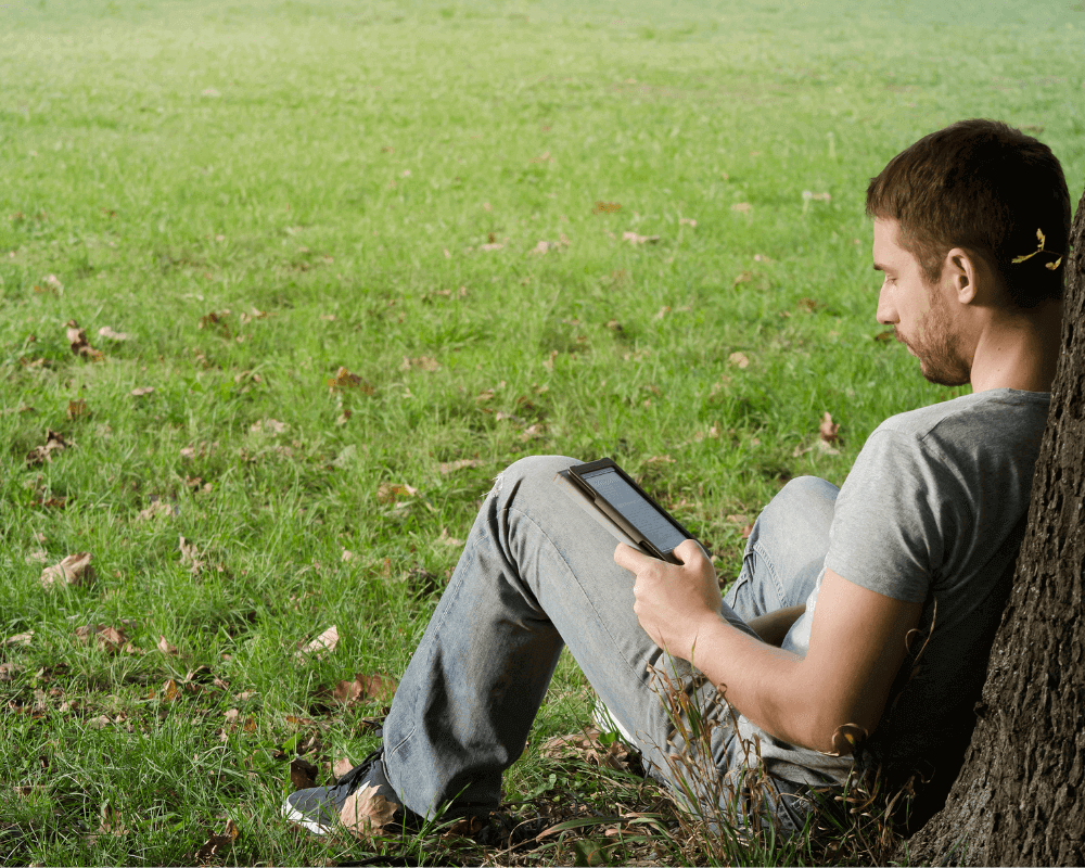 木によしかかり、Kindleで本を読んでいる男性